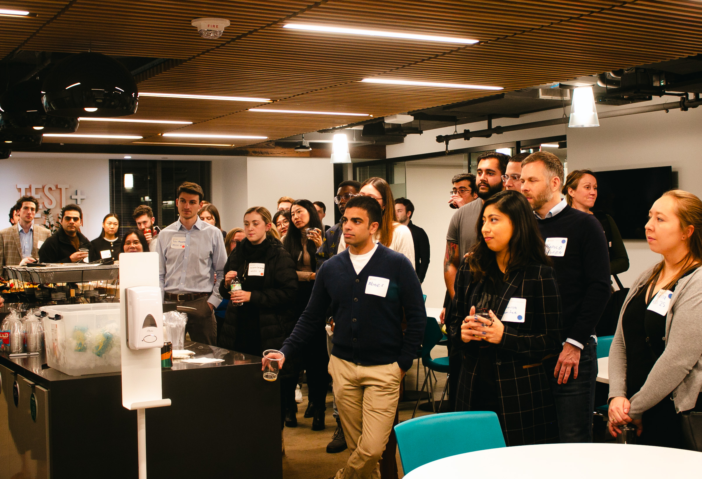 Many students stand together during a social event, facing the speaker.