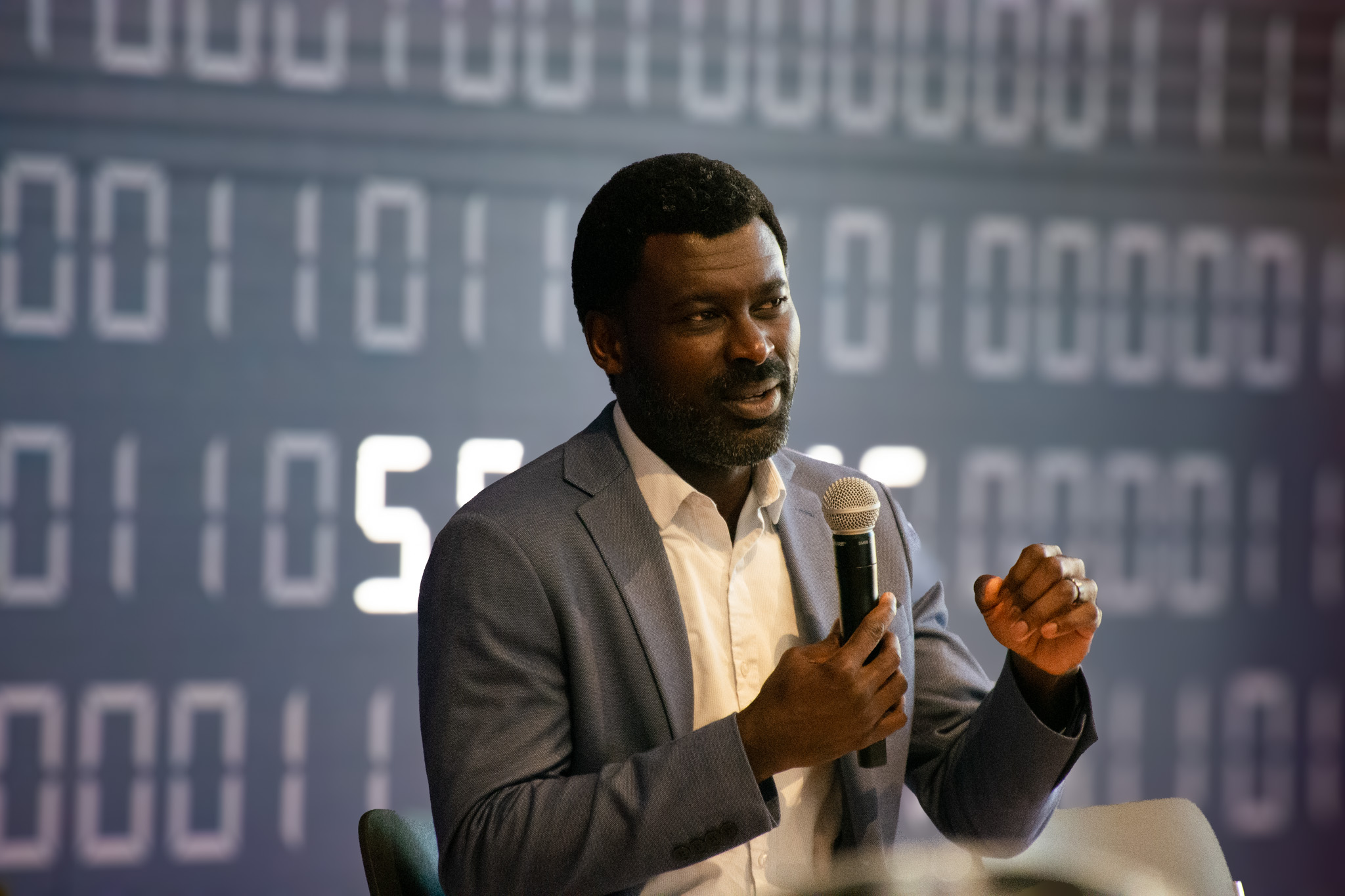 Prince Afriyie holds a microphone, addressing the audience in front of a dark blue media wall