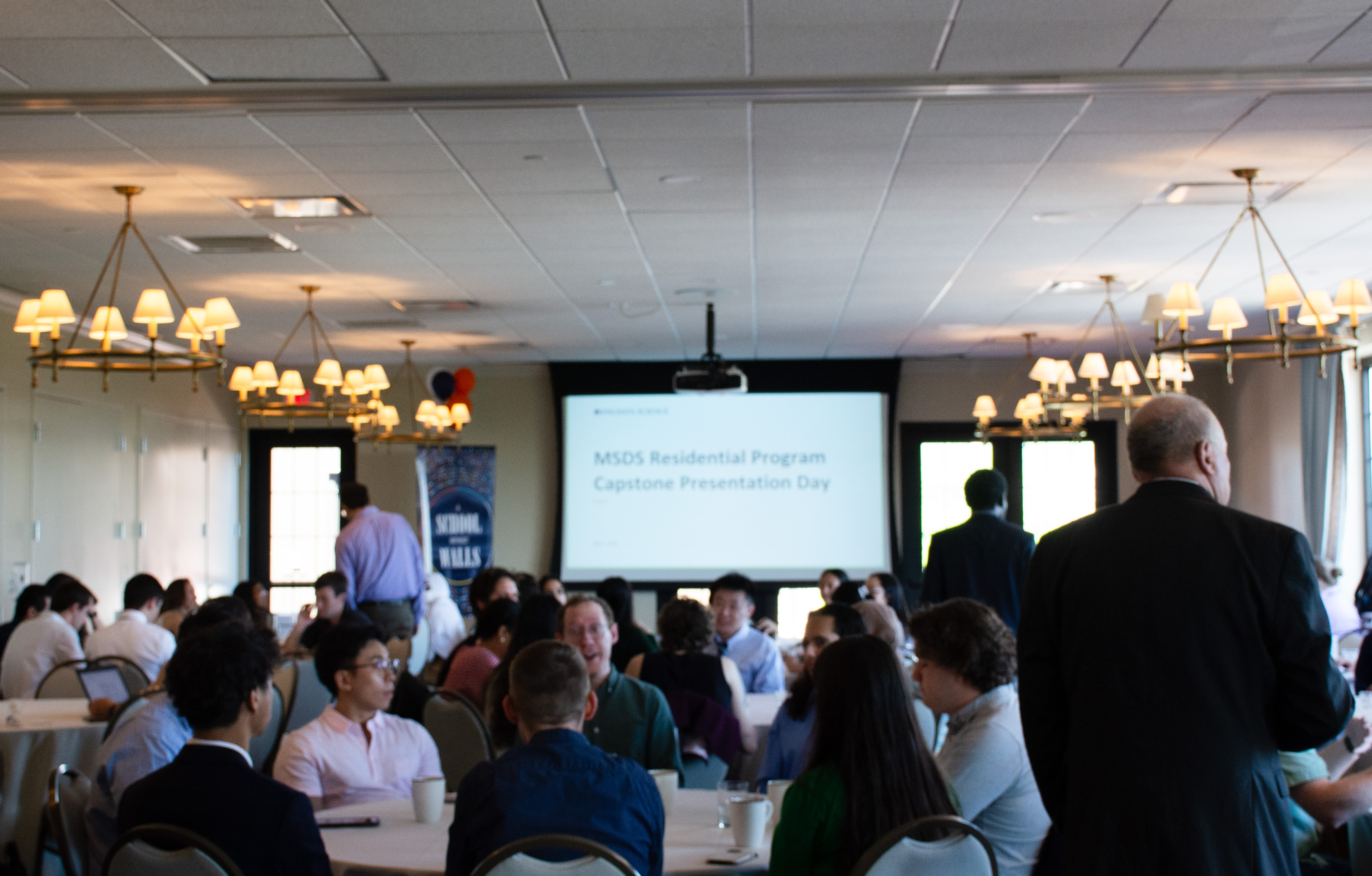 Students sit in circular tables and an event room, chatting prior to capstone presentations.