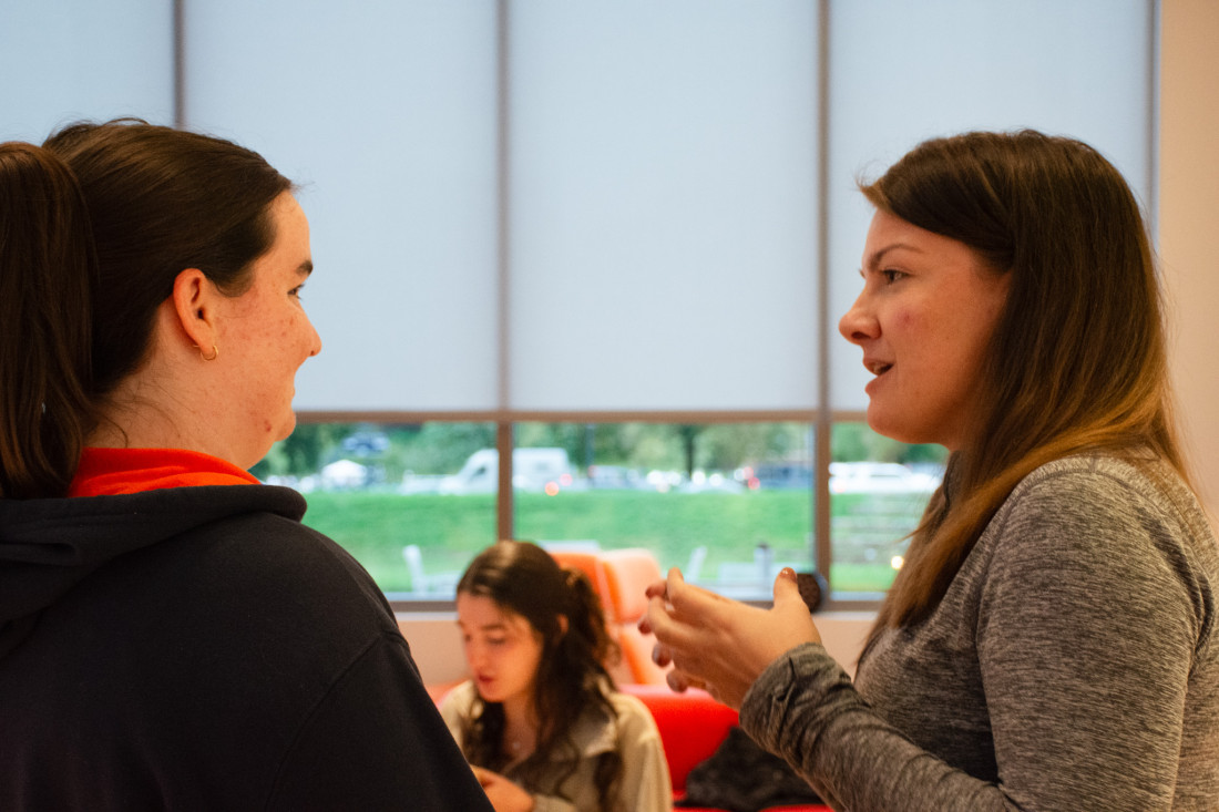 Sarah Dodge converses with a student in front of large windows with the blinds pulled halfway down inside of the School of Data Science.