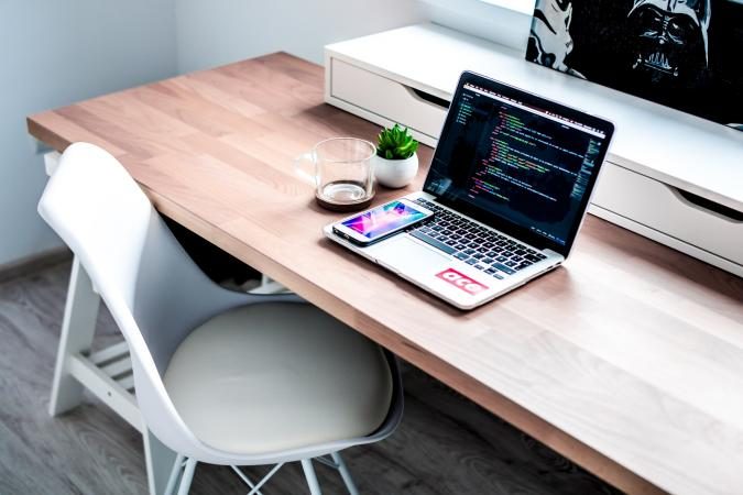 A minimalist workspace features a laptop displaying code, a smartphone, small plant, and a mug on a wooden desk. 