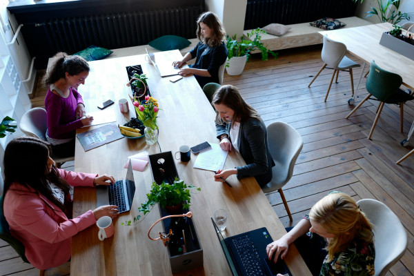 Five women sit at an office table, working together. 