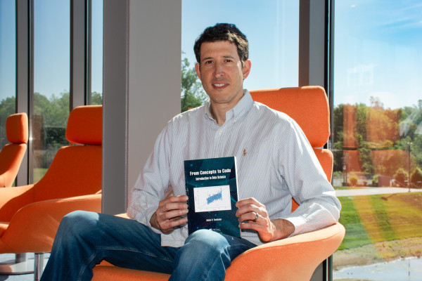 Adam Tashman wearing a blue shirt sits in an orange chair by a large window inside of the School of Data Science, holding a book titled "From Concept to Code." 