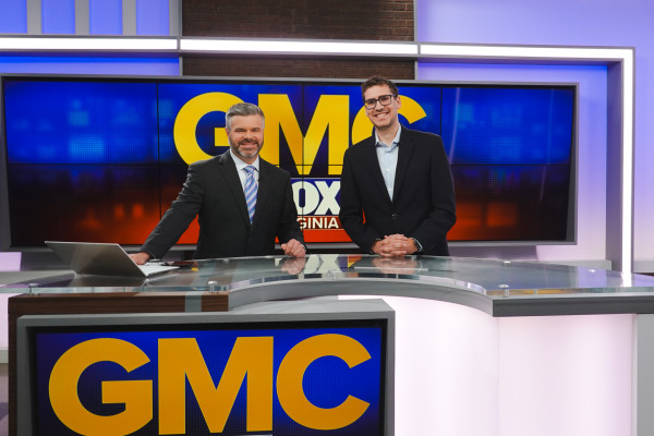 Jonathan stands with one news anchor, posing in front of the GMC logo.
