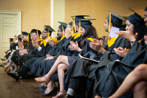 2019 MSDS Graduates in caps and gowns sit and applaud in a brightly lit room. They appear joyful and proud, capturing a celebratory, academic atmosphere.