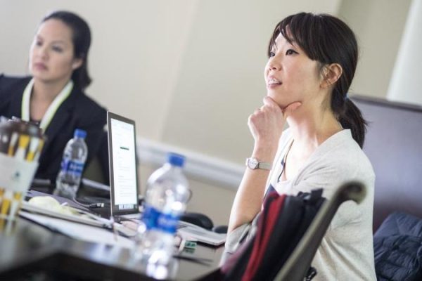 Two women in a meeting room focus on a laptop screen, surrounded by water bottles and papers. One woman sits thoughtfully, hand on chin. 