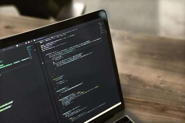 Laptop displaying code on a wooden desk, suggesting programming or software development. The screen's dark theme contrasts with the workspace.