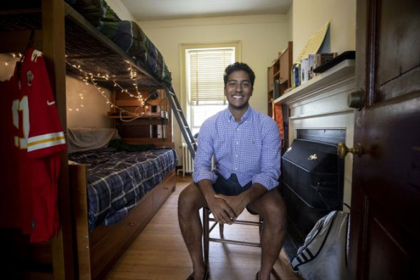 Ashwanth Samuel on the Lawn smiles while sitting on a chair in a cozy dorm room. The room features a bunk bed with string lights, a red sports jersey, and a fireplace.