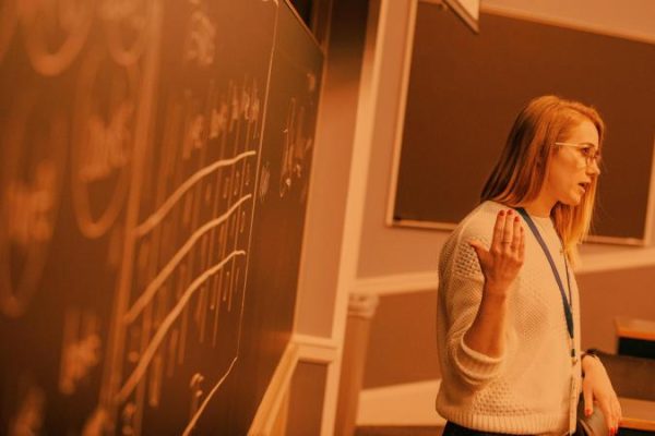 A woman in a white sweater gestures while speaking beside a chalkboard filled with diagrams. The classroom setting conveys focus and engagement.