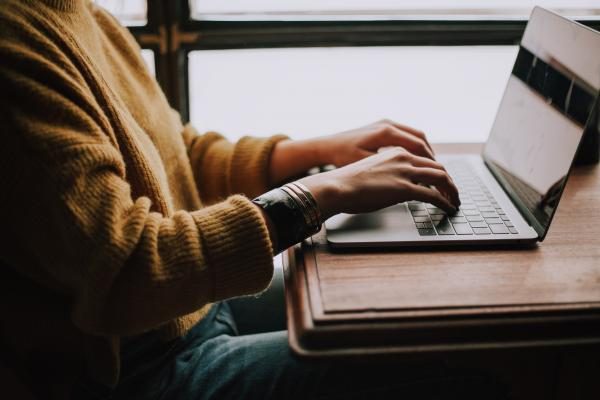 A person in a yellow sweater types on a laptop at a wooden desk, natural light from a window.