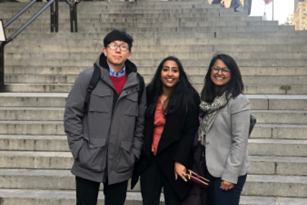 Logan Lee, Sudeepti Surapaneni, and Sana Syed stand smiling on wide stone steps in front of a historic building. They seem relaxed, with people and an American flag in the background.