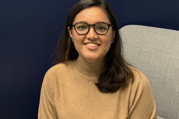Charu Rawat, with long dark hair and glasses smiles while sitting on a gray chair. She is wearing a beige sweater. The word "London" is on the navy blue wall behind her.