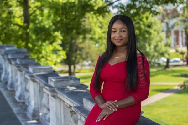 Oretha Domfeh in a red lace dress sits on a stone railing with a garden in the background. She smiles softly, projecting confidence and calm under the sunlight.