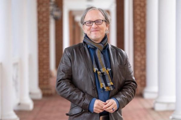 Eric Tassone stands in a corridor with brick walls and white columns, wearing glasses, a leather jacket, and a colorful scarf, conveying a relaxed and confident demeanor.