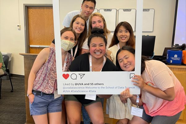 MSDS Residential Class of 2023, a group of eight diverse people smiling and posing together inside a large Instagram frame prop, labeled "School of Data Science," in a classroom setting.