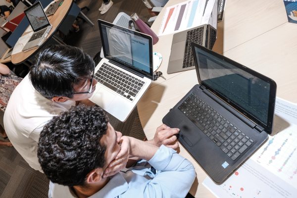 Two people collaborate at a desk with laptops, viewing data and charts. The environment is focused and analytical, suggesting teamwork and problem-solving.