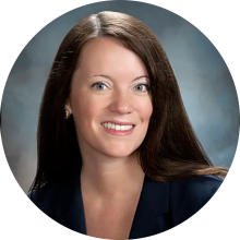 Sarah smiling wearing a blue blazer in front of a bluish gray backdrop.